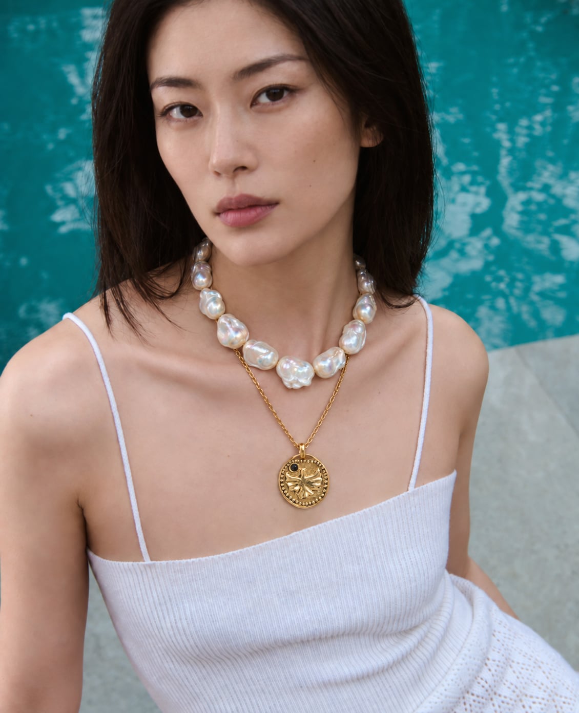 Model wearing layered necklaces with white beads and a round gold-tone pendant against a turquoise background