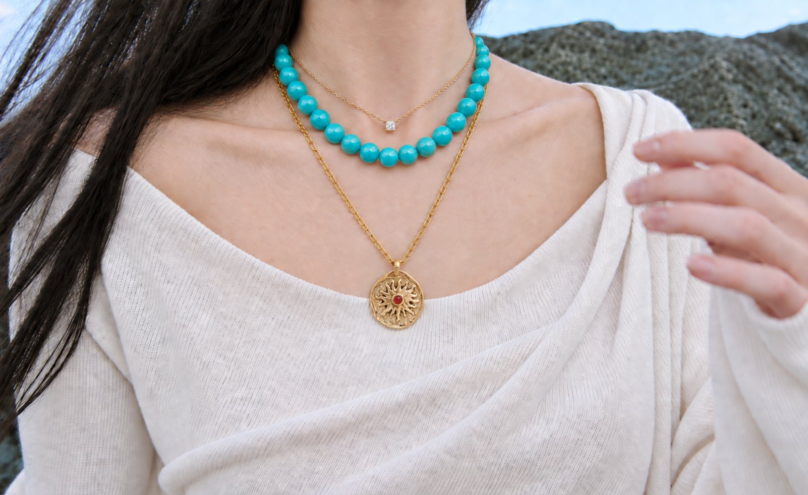Turquoise-blue bead necklace layered with gold-tone necklaces and a sun pendant over a white knit top